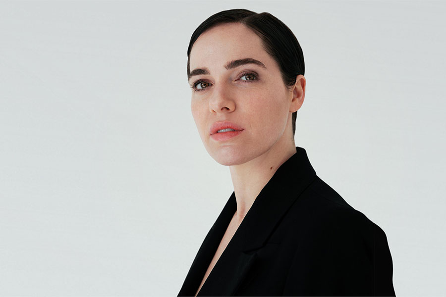 Professional headshot of Austrian actress Verena Altenberger with slicked-back dark hair, wearing a black blazer against a plain white background.