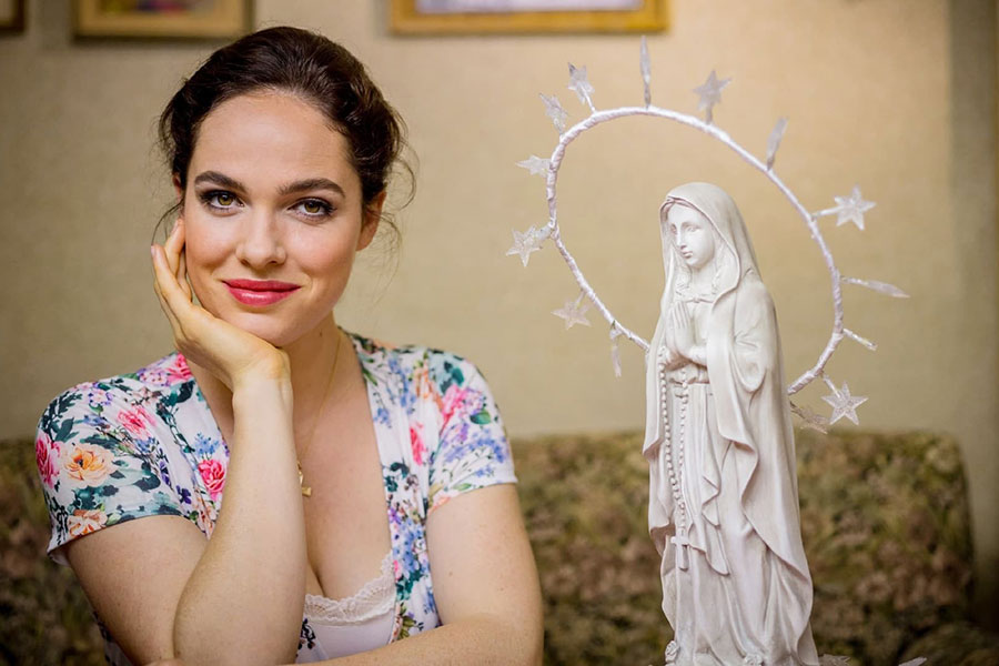 Verena Altenberger in character for the German comedy series 'Magda macht das schon!', smiling while seated next to a statue of the Virgin Mary.