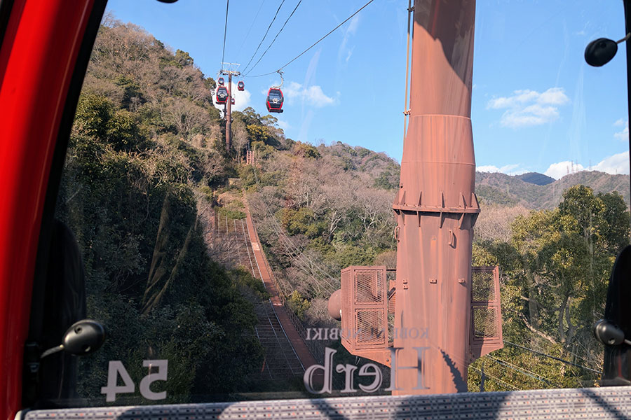 View from inside a gondola on the Kobe Nunobiki Ropeway ascending Mount Rokko, with red cable cars and forested hills in the background