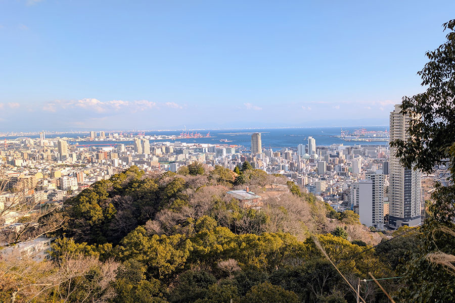 Expansive view of Kobe skyline and harbor from the mountains, with city buildings, port facilities, and Osaka Bay under a clear blue sky