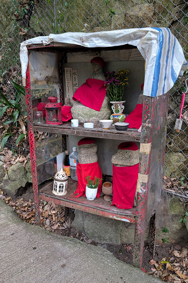 Small roadside shrine near Nunobiki Waterfalls in Kobe, Japan, featuring red-cloaked stone statues, offerings, and protective coverings