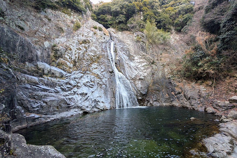 Wide landscape view of On-taki Waterfall near Shin-Kobe, Japan, with a serene basin at the base and steep rocky cliffs in the background