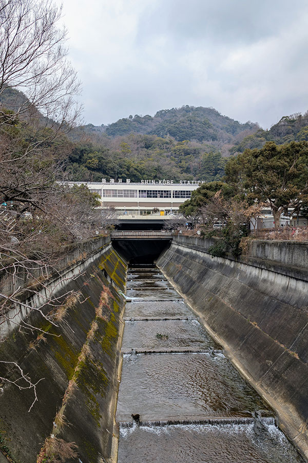 View of Shin-Kobe Station from across a concrete river channel, framed by forested hills under a cloudy sky in Kobe, Japan