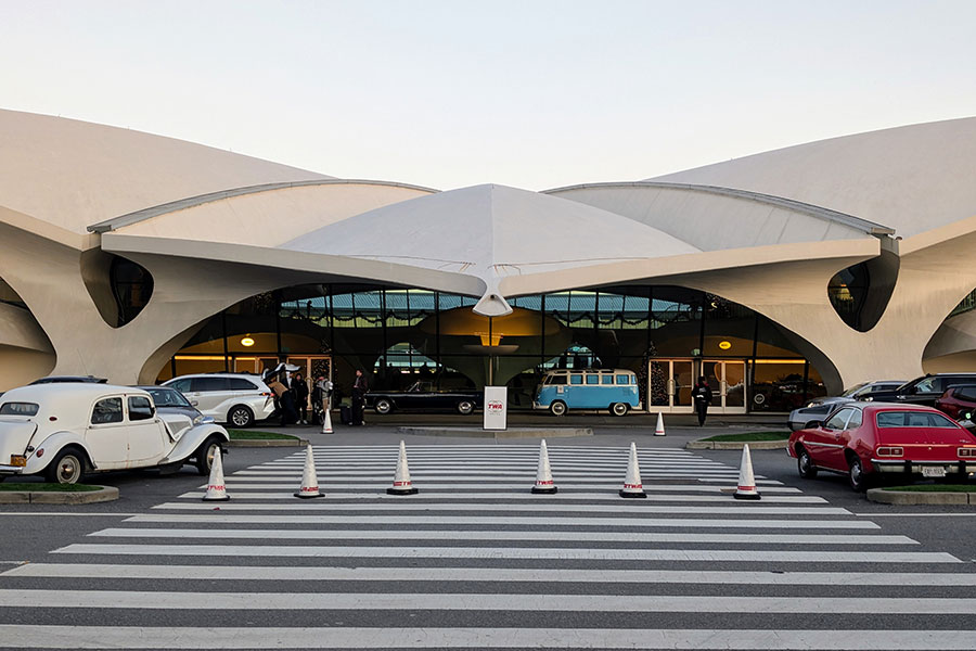 Front exterior view of the former TWA Flight Center, now the TWA Hotel, with a crosswalk leading to its entrance and classic vehicles on display.
