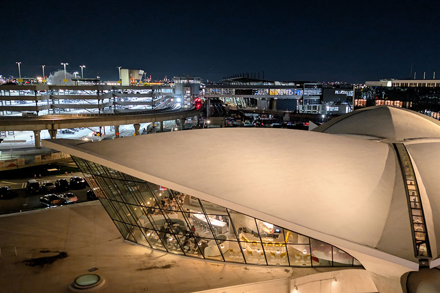 The beautifully lit exterior of the TWA Hotel at night, contrasting with the busy activity of JFK Airport seen in the distance.