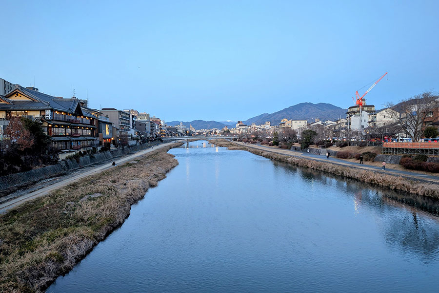 The Kamo River in Kyoto at dusk during winter, with traditional buildings on the left, mountains in the distance, and a clear blue sky