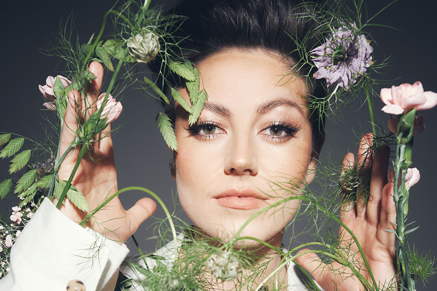 Artistic portrait of German comedian and impressionist Tahnee looking confidently at the camera, wearing a white blazer with flowers and green foliage framing her face.