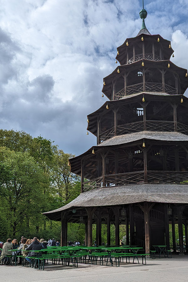The iconic wooden Chinese Tower (Chinesischer Turm) pagoda in Munich's Englischer Garten, with green beer garden tables and benches in the foreground under a cloudy sky.