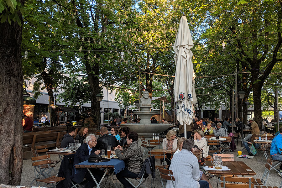 Patrons sitting at wooden tables under large chestnut trees at the Paulaner am Nockherberg Biergarten in Munich, with a fountain visible in the background.