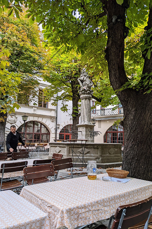 The shady courtyard Biergarten of the Hofbräuhaus in Munich, with tables set under large chestnut trees near the central statue.