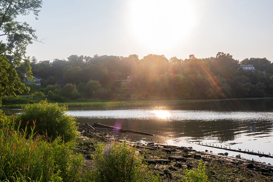 Golden hour sunset over the Hudson River, with the bright sun reflecting on the water and silhouetting the trees on the far shore.