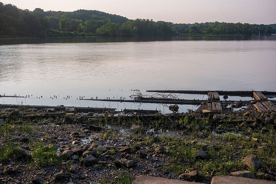 The calm, quiet Hudson River at dusk, with the decaying wooden pilings of an old pier emerging from the water in the foreground.