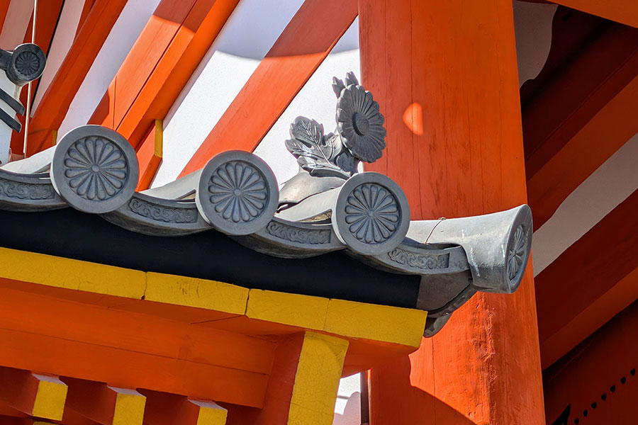A close-up of grey Japanese roof tiles featuring the Imperial Chrysanthemum Seal on a bright orange wooden structure at the Kyoto Imperial Palace.