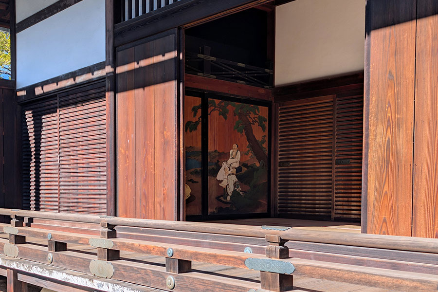 A view across a wooden veranda into a palace room, revealing a beautifully painted fusuma sliding screen depicting traditional figures in a landscape.
