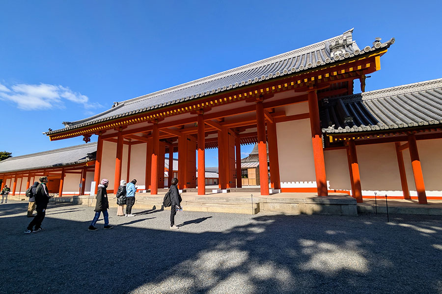 Visitors walking towards the Jomeimon Gate at the Kyoto Imperial Palace, a grand entrance with bright vermilion pillars, white walls, and a traditional tiled roof.