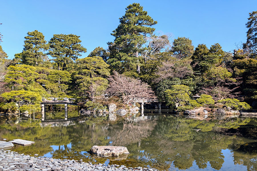The large pond of the Oikeniwa Garden at the Kyoto Imperial Palace, with a clear, mirror-like reflection of the lush pine trees and a distant bridge on its calm surface.