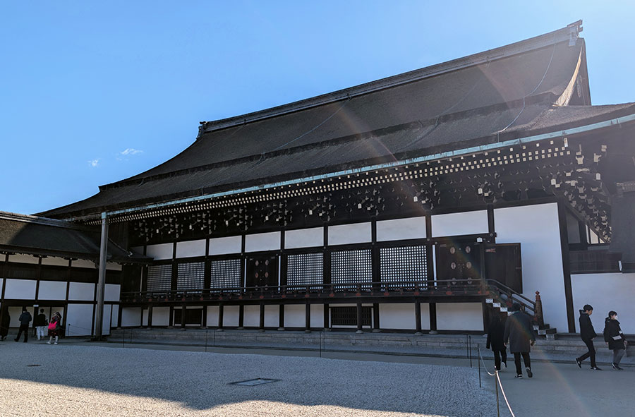 The grand exterior of the Shishinden Hall at the Kyoto Imperial Palace, showing its vast cypress bark roof, white walls with dark timber frames, and the wide gravel courtyard in front.