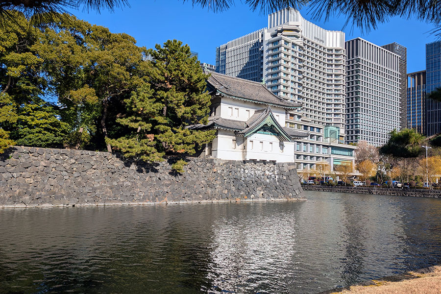 The Sakurada-Tatsumi Yagura, a two-story corner keep at the Tokyo Imperial Palace, sits atop a stone wall next to the moat, with modern skyscrapers rising behind it.