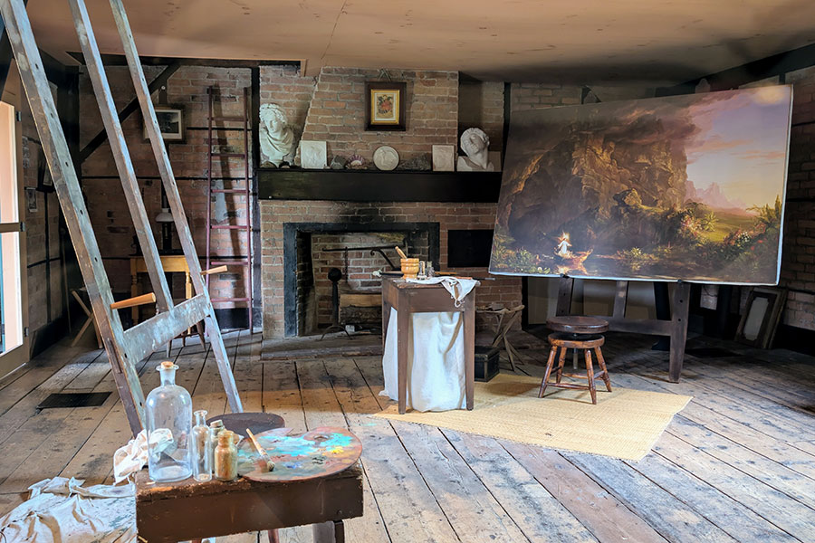 Inside the historic art studio at the Thomas Cole National Historic Site, with an easel and a large Hudson River School painting next to the fireplace.