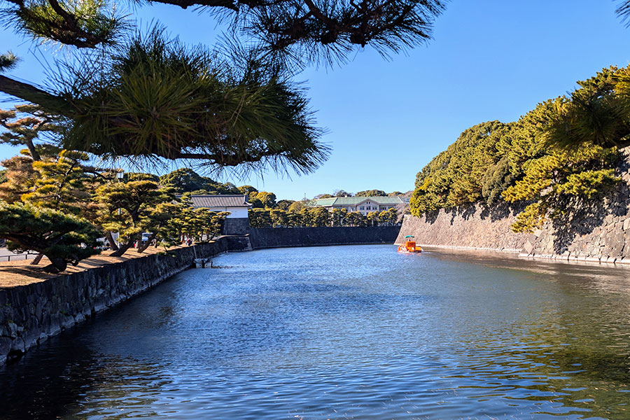 A view down the wide moat of the Tokyo Imperial Palace, lined with massive stone walls and manicured Japanese black pine trees under a clear blue sky.