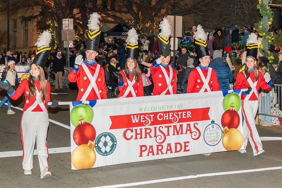 A group of smiling marching band members in festive red uniforms carry a large welcome banner to kick off the West Chester holiday parade at night.