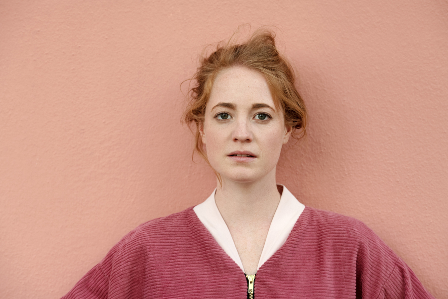 Portrait of German actress Leonie Benesch wearing a maroon corduroy jacket against a textured pink wall