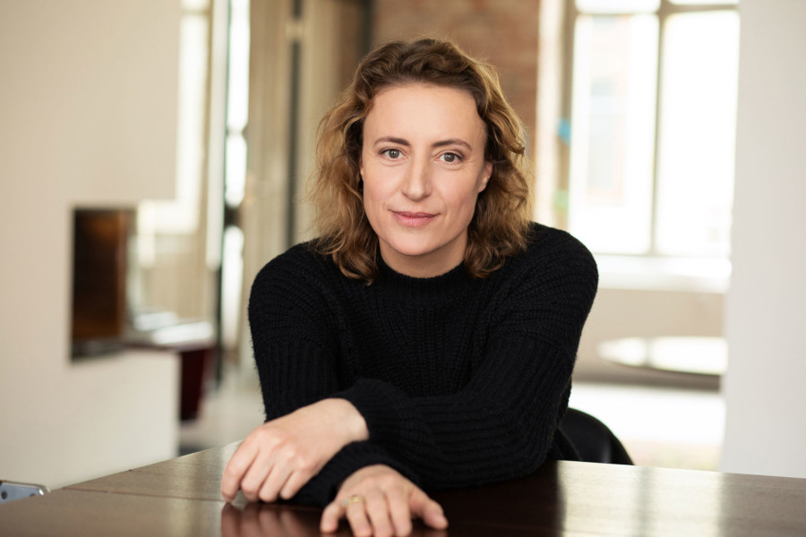 Portrait of German actress Maren Eggert smiling while sitting at a dark wood table, wearing a black ribbed sweater.