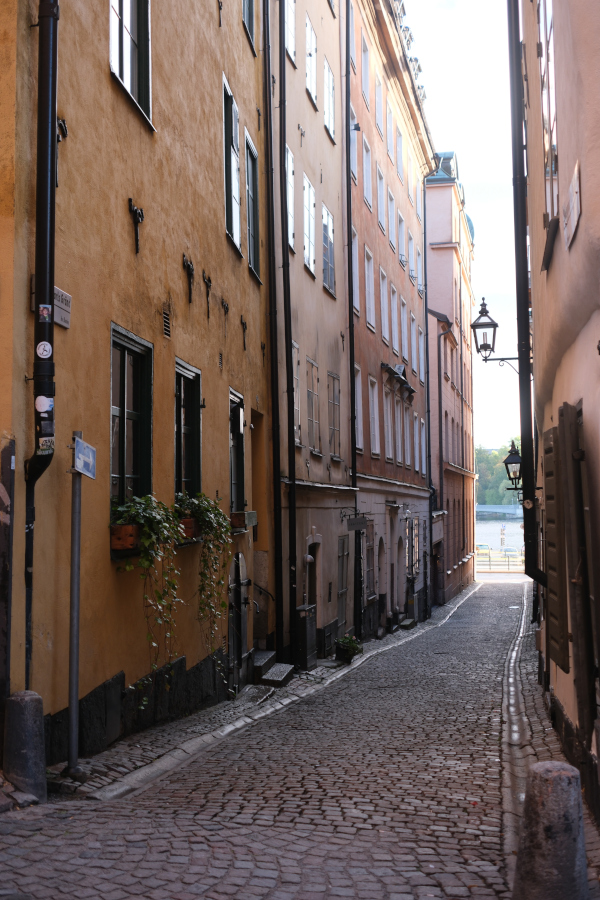 A narrow, historic cobblestone alleyway in Gamla Stan, Stockholm's Old Town, lined with warm ochre-colored buildings and traditional wrought-iron street lamps.
