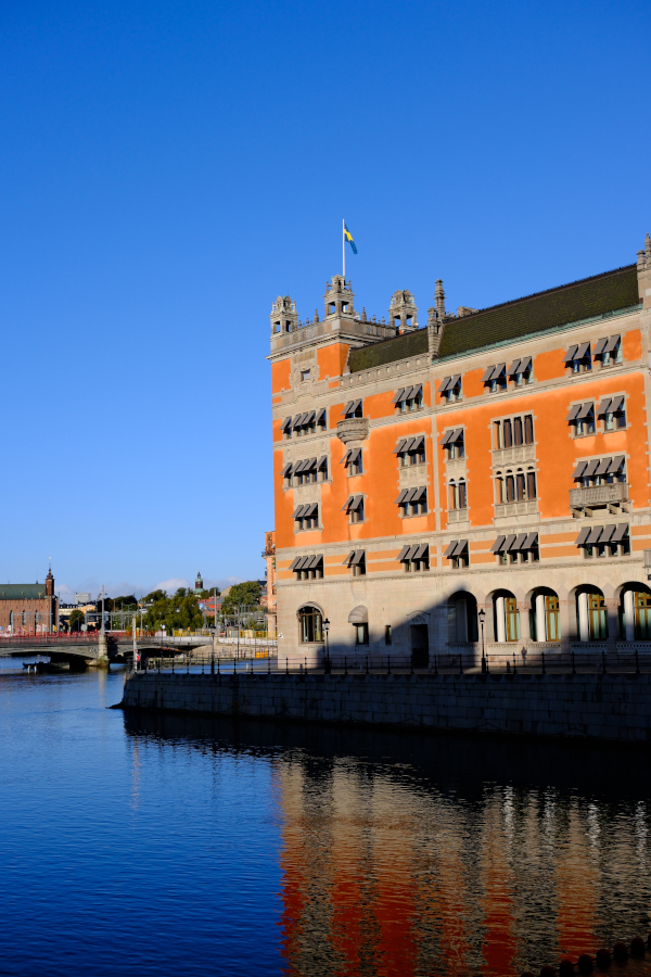 The vibrant orange Rosenbad building reflecting in the calm blue water of the Stockholm harbor under a clear sky on a crisp autumn morning.