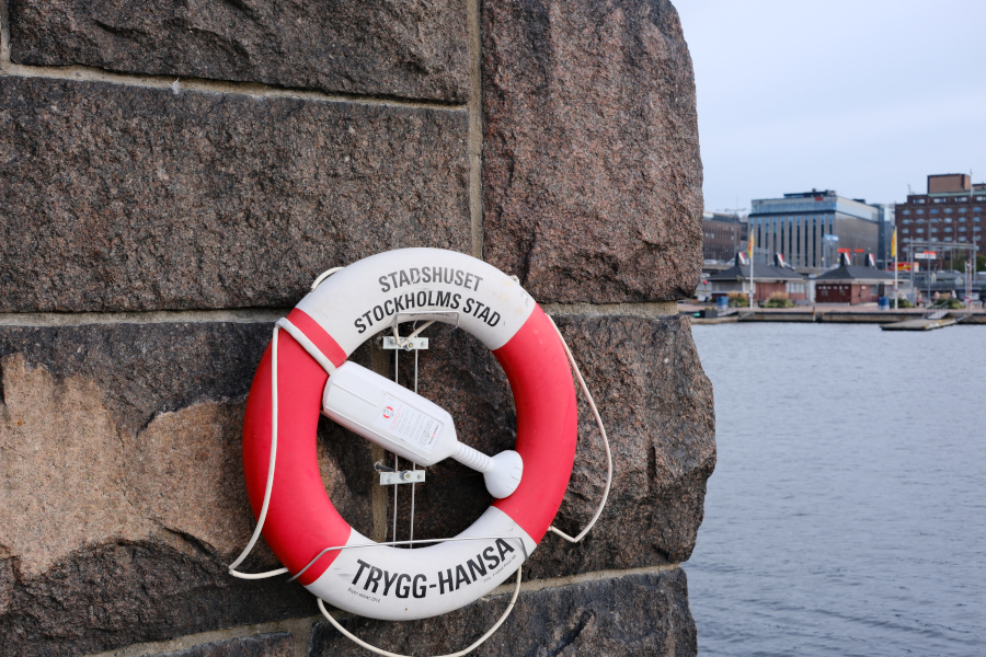 A classic red and white Trygg-Hansa lifebuoy mounted on a textured stone wall overlooking the Stockholm harbor near City Hall.