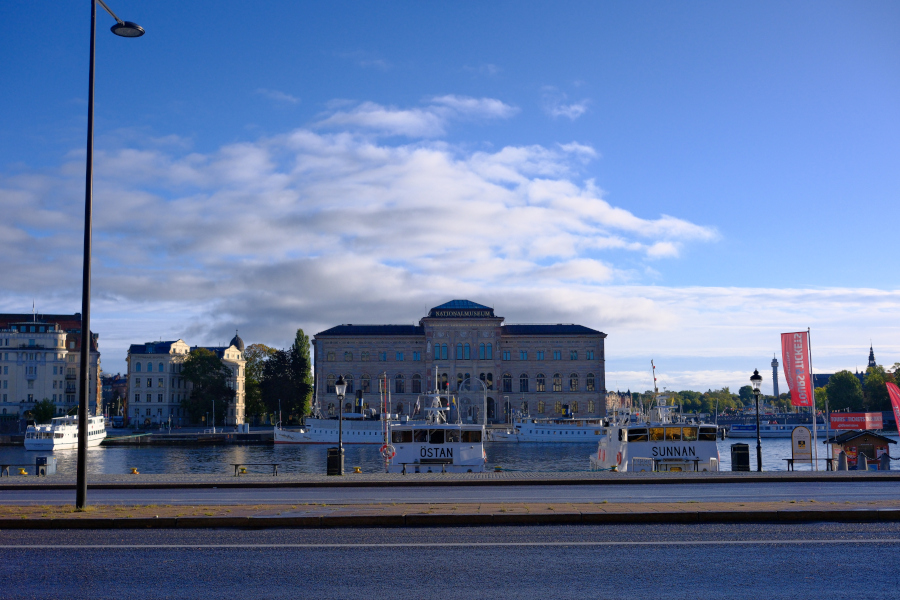 Wide view of the Nationalmuseum of Sweden across the water, with white passenger boats docked under a blue sky with soft white clouds.