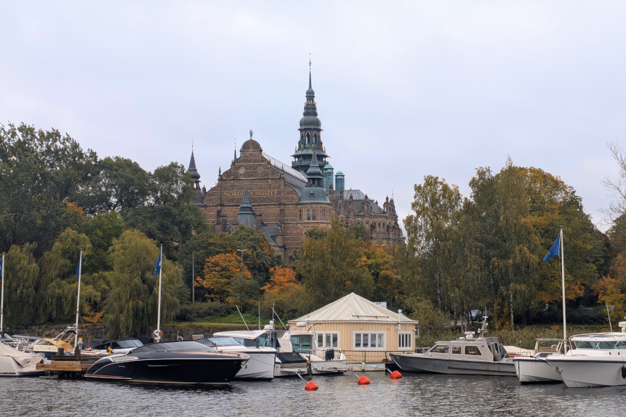 View of the grand Nordiska museet building rising above autumn foliage and white motorboats docked in the harbor on a cloudy day.
