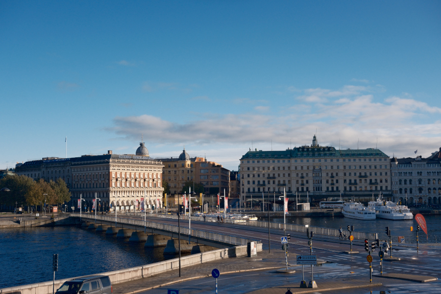 A wide view of the Norrström waterfront in Stockholm, featuring the grand historic architecture of the Royal Swedish Opera area and a stone bridge crossing the water.