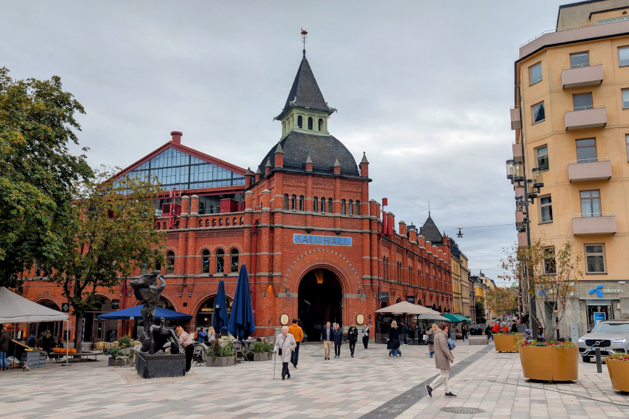 The iconic red-brick facade and turret of the Östermalm Saluhall food market, with pedestrians walking through the square on a typical Stockholm afternoon.