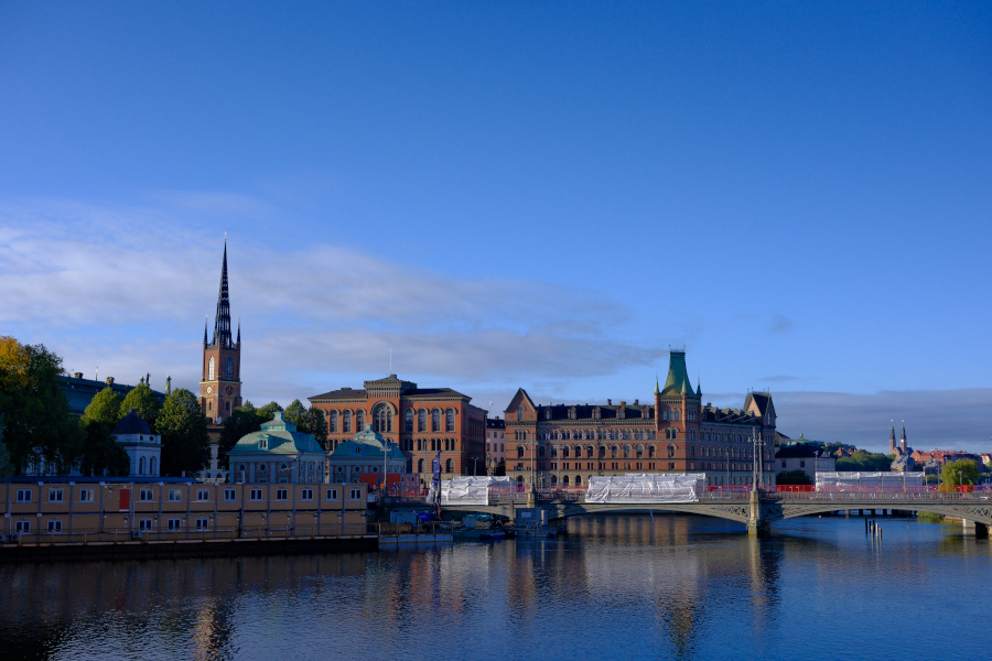 The striking latticework spire of Riddarholmen Church and historic brick buildings overlooking the serene, blue waters of the Stockholm harbor at sunrise.