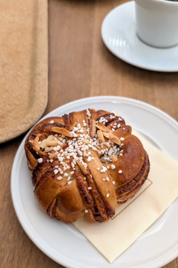 Swedish cinnamon bun (kanelbulle) topped with pearl sugar and sliced almonds on a plate beside a cup of coffee during a traditional fika