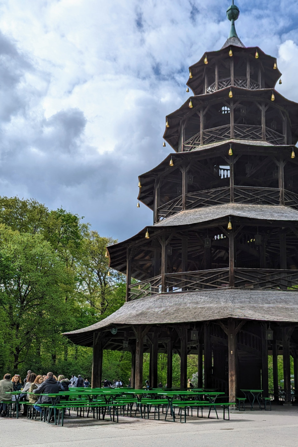 The iconic wooden Chinese Tower and beer garden seating in Munich's English Garden under cloudy skies.
