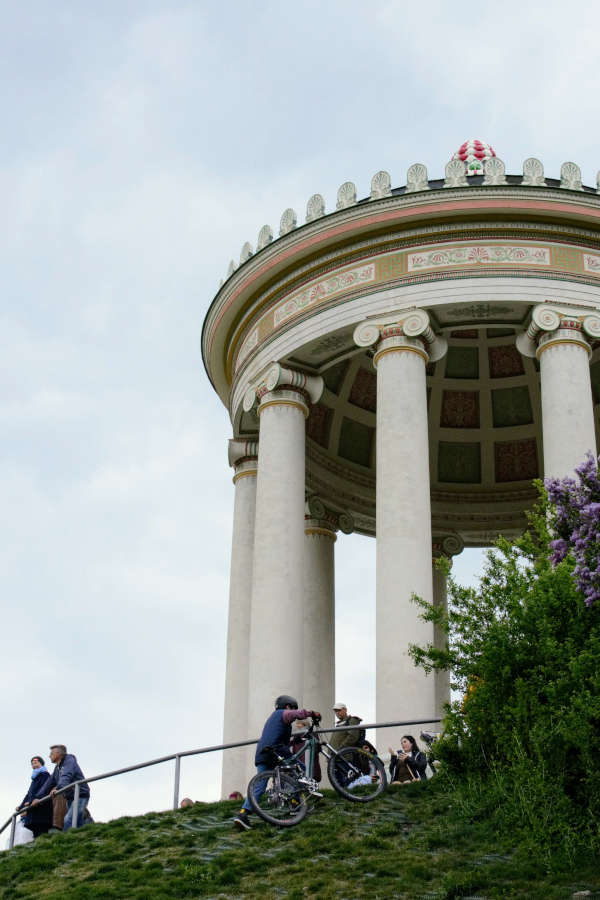 Visitors and a cyclist climbing the grassy hill toward the Monopteros temple in Munich.