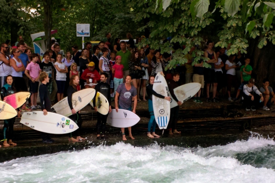 Surfers holding surfboards lined up along the Eisbachwelle wave in Munich's English Garden.