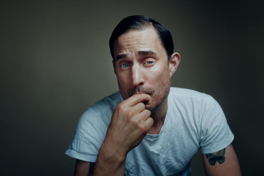 A close-up portrait of German actor Clemens Schick wearing a white t-shirt and thoughtfully holding a finger to his lips, with a butterfly tattoo visible on his arm.