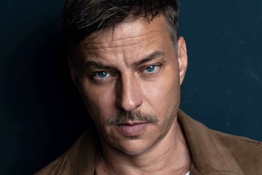 Close-up portrait of German actor Tom Wlaschiha with a mustache, looking directly at the camera against a dark background.